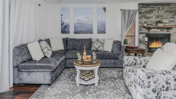 A living room with a grey sectional sofa, a coffee table with decorative items, a fireplace, and a view of mountains through the windows. - Aspen Run Condo Unit 29A | Great Views & Location! (Ruidoso)