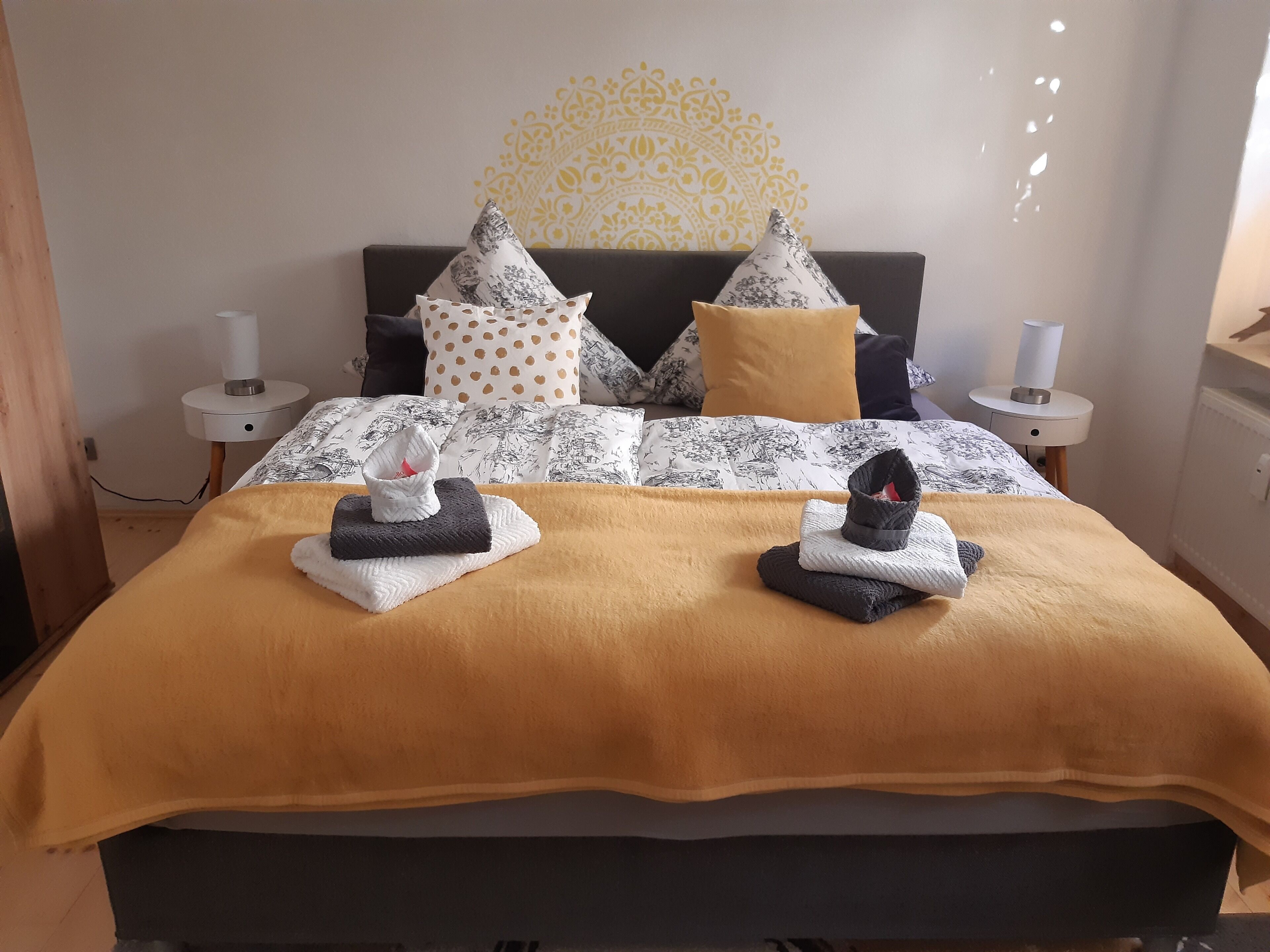Ein ordentlich bezogenes Bett mit einer gemusterten Tagesdecke, zwei Kissen und zwei gefalteten Handtüchern.