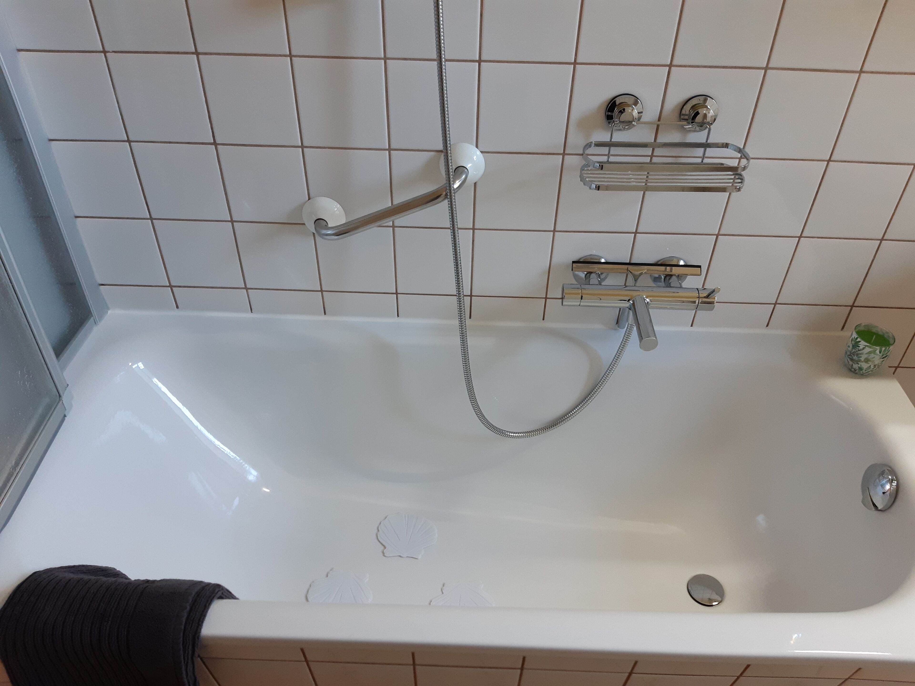 A white bathtub with a handheld showerhead, a glass shelf with toiletries, and a small green candle on the wall.