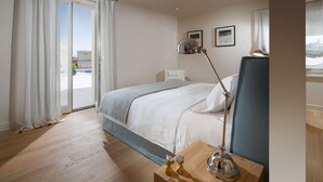 Hotellihuone, jossa on sänky, yöpöytä lampun kanssa, ikkuna verhoilla ja näkymä ulos.