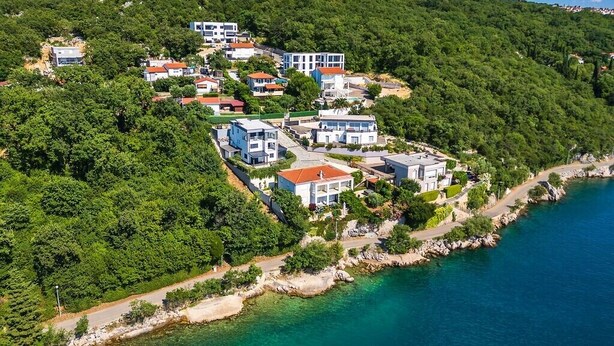 An aerial view of a coastal village with buildings on a hillside, a clear blue sea, and a rocky shoreline.