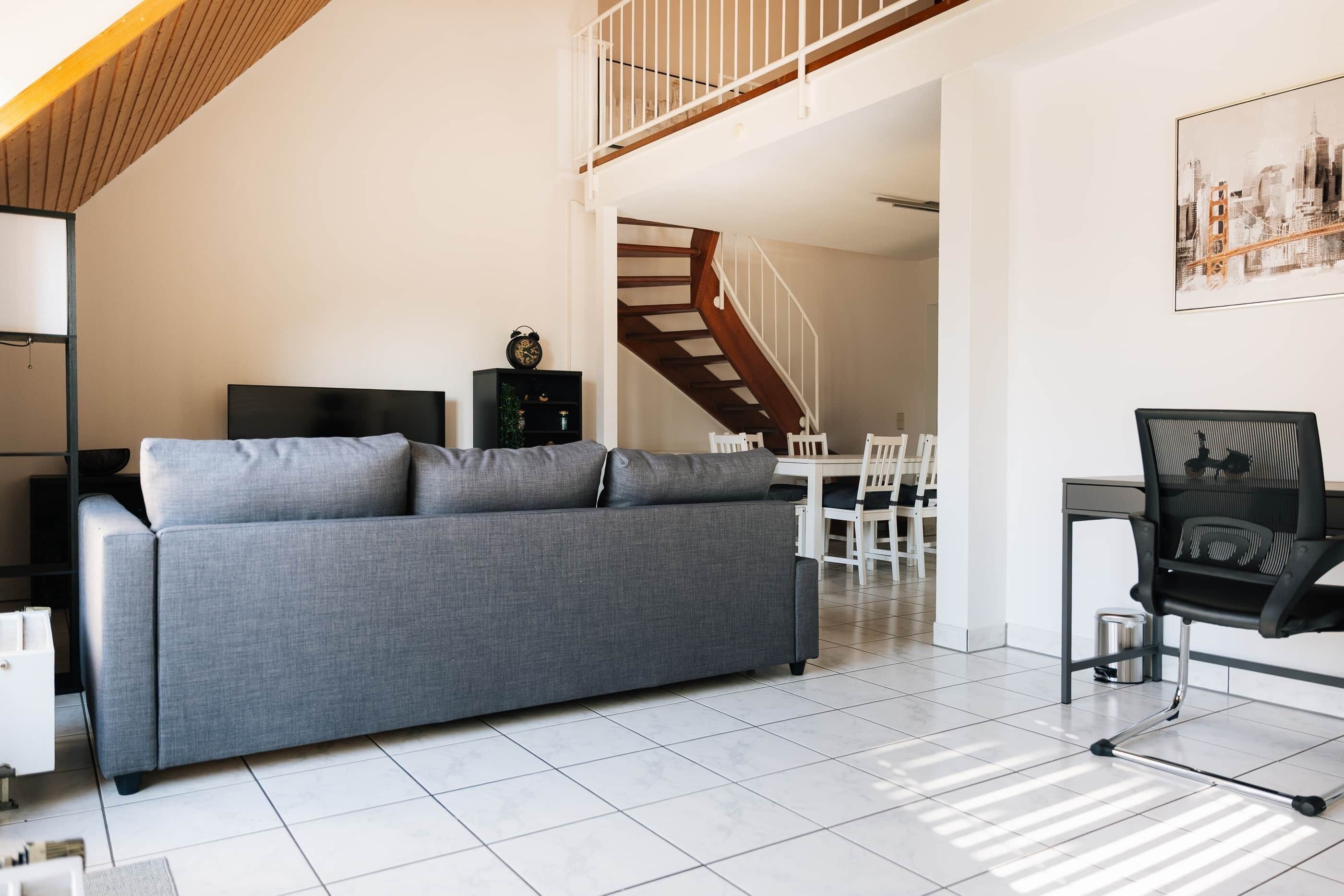 Ein modernes Wohnzimmer mit grauem Sofa, schwarzem Bürostuhl, Esstisch mit Stühlen und einer Treppe, die zu einer oberen Ebene führt.