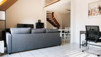 Una sala de estar moderna con un sofá gris, una silla de oficina negra, una mesa de comedor con sillas y una escalera que conduce a un nivel superior.