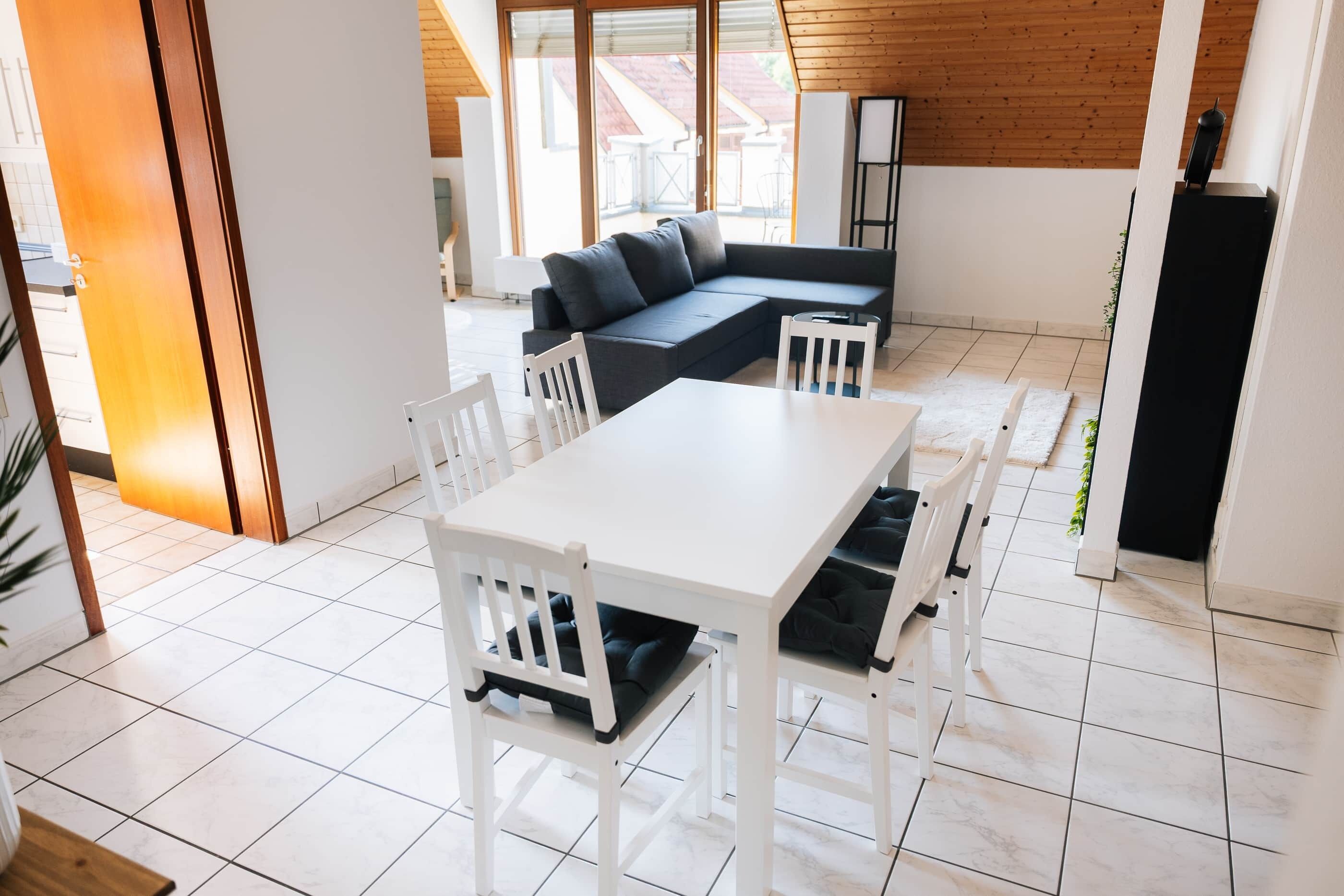 Ein modernes Interieur mit einem weißen Esstisch, weißen Stühlen, einem dunklen Sofa und einem weißen Schrank.