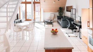 Eine moderne Küche mit einer zentralen Kochinsel, einem Essbereich mit weißer Möbel, einer Treppe und einem Wohnbereich mit Sofa und Pflanze.