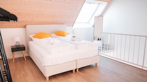Ein Schlafzimmer mit schrägen Wänden, einem Bett mit weißer Bettwäsche und orangen Kissen, einem kleinen Nachttisch mit Lampe und einer weißen Geländer.