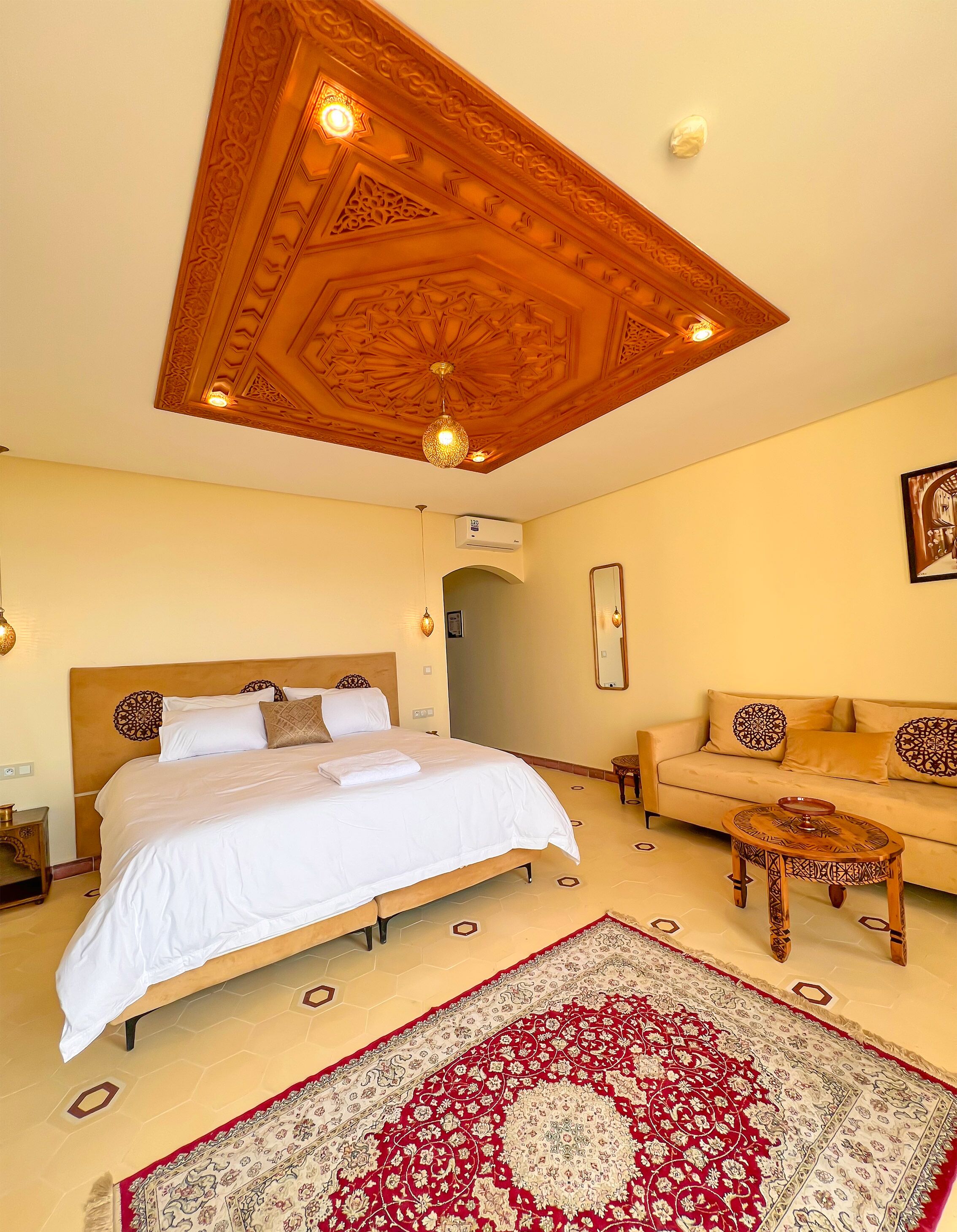 A bedroom with a large bed, a sofa, a patterned rug, and a decorative ceiling.