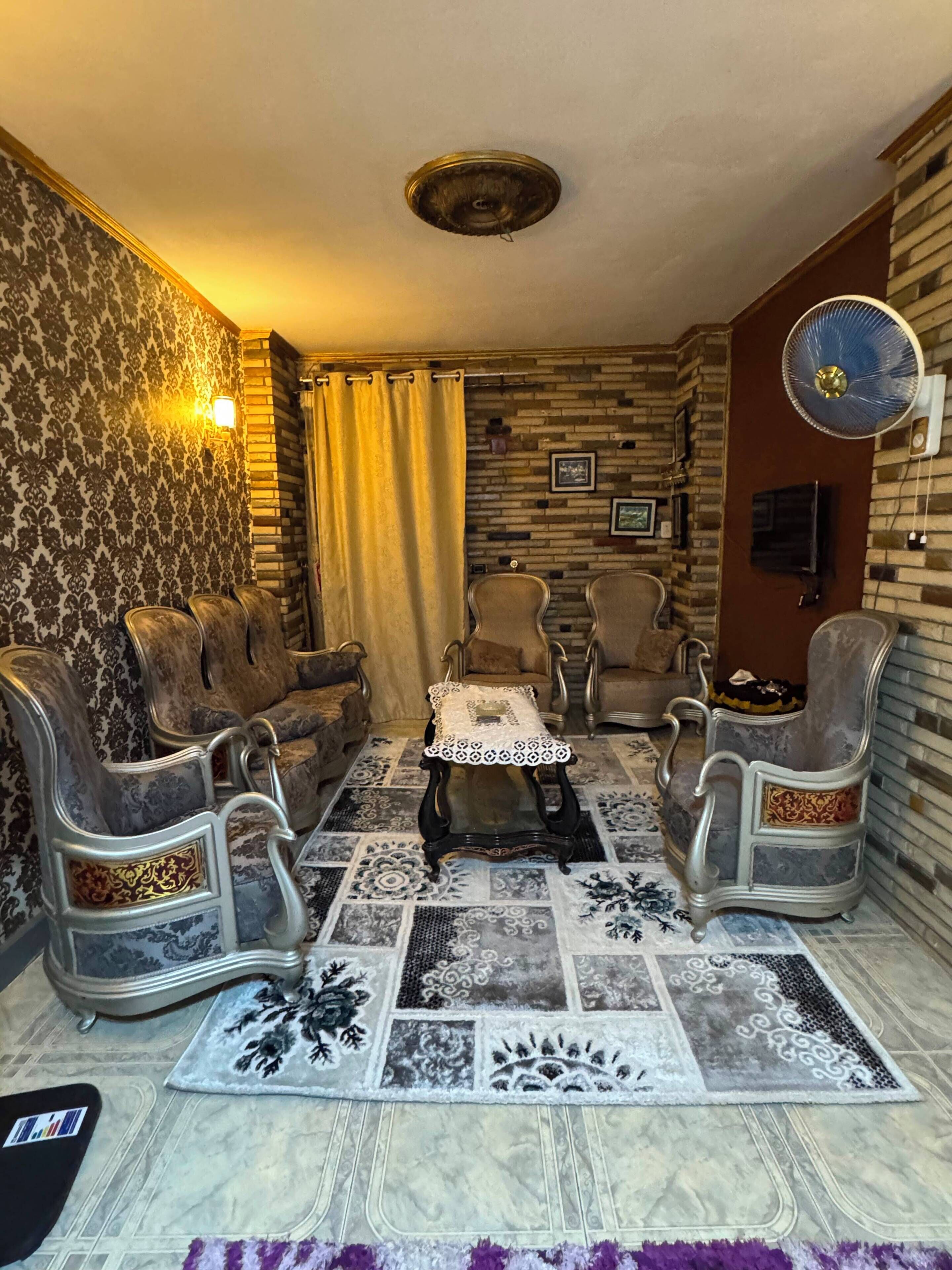 Ein Zimmer mit gemustertem Teppich, einer Sofagarnitur, einem kleinen Tisch mit dekorativer Abdeckung, einem Wandventilator und einer Ziegelwand.
