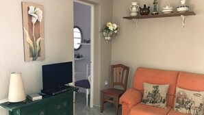 Ein Wohnzimmer mit einem Fernseher auf einer grünen Kommode, einem orangefarbenen Sofa mit Zierkissen, einem Holzstuhl und einer Wandregal mit Dekorationen.