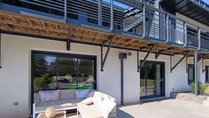 A modern outdoor seating area with a sofa and chairs, a wooden table, and a large sliding glass door. - Family Retreat with Terrace and Pool Access (Jeanménil)