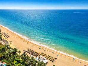Ein Strand mit einer Reihe von Liegestühlen und Sonnenschirmen, einem klaren, blauen Meer und einer fernen Küstenlinie.