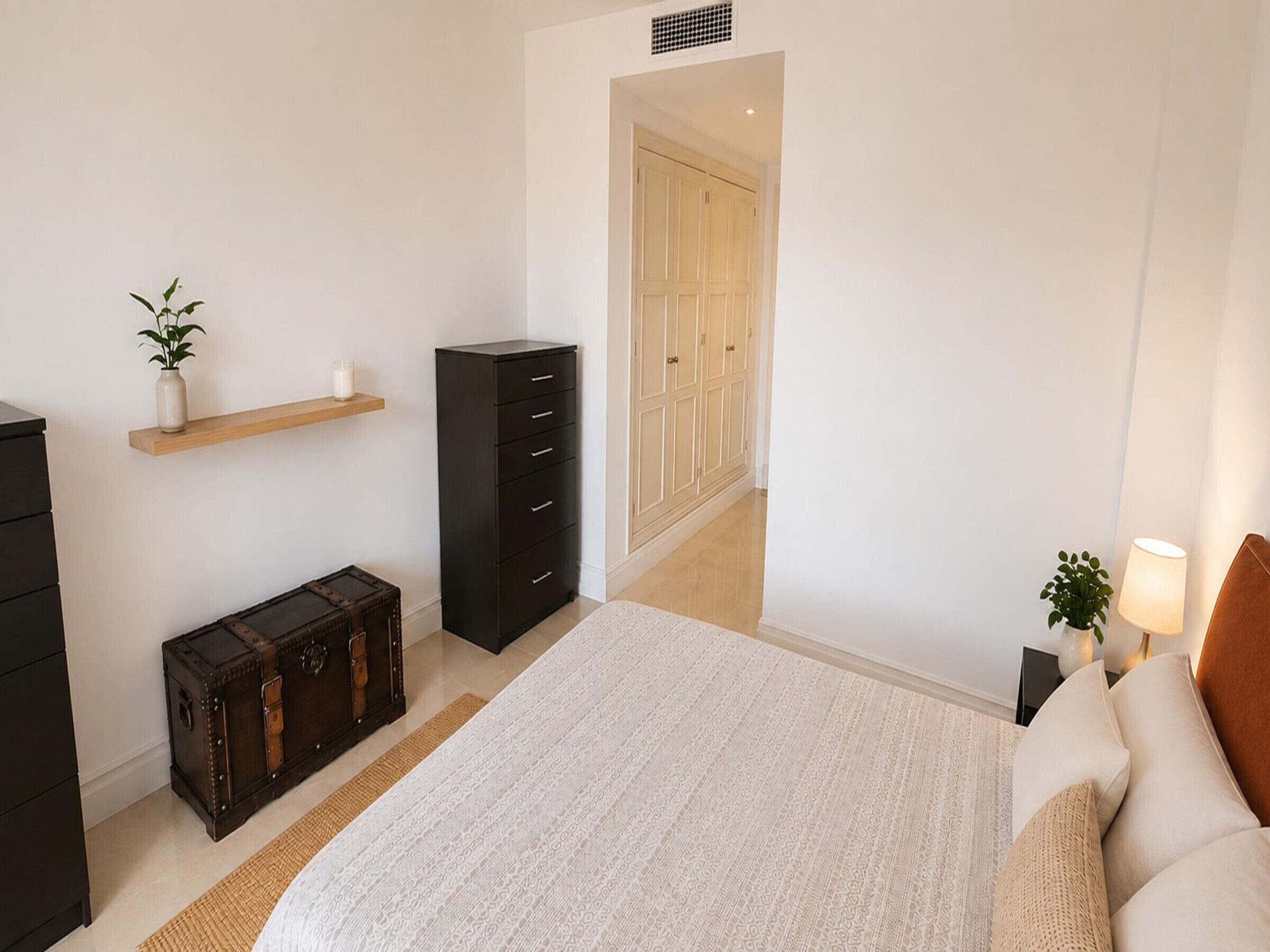 Une chambre à coucher comprenant un lit, une commode, une table de chevet, une lampe, une plante en pot, une étagère en bois et une armoire.