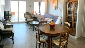 Ein geräumiger Wohnbereich mit Sofa, Sesseln, einem Esstisch mit Obstschale, einem Fernseher und einer Vitrine.