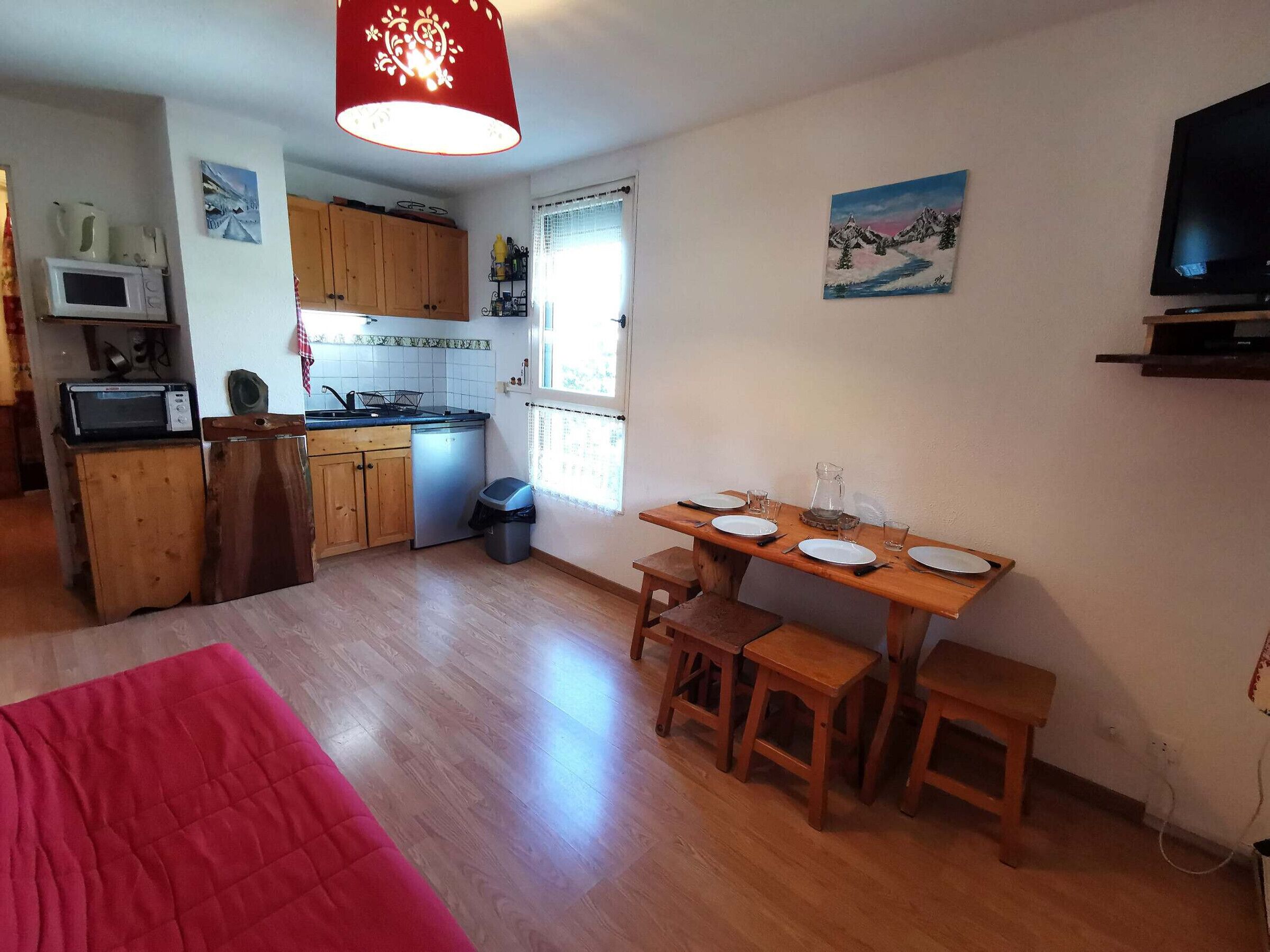 Eine kleine Küche mit Holzausstattung, einer Mikrowelle und einem Spülbecken. Es gibt einen Essbereich mit Tisch für zwei Personen, ein rotes Bett im Vordergrund und ein Bild an der Wand.