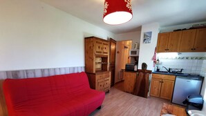Ett kompakt bostadsutrymme med en röd soffa, träskåp, en liten köksdel och en matplats med ett bord dukat för två personer.