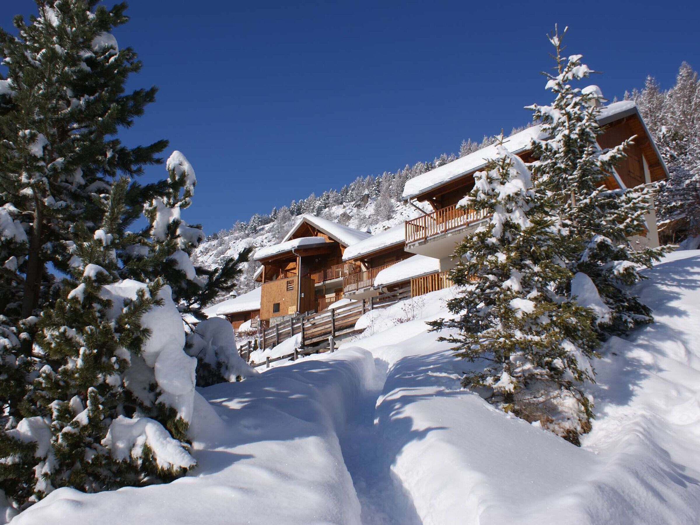 A snow-covered lodge with multiple wooden balconies, surrounded by snow-laden trees and a clear blue sky.