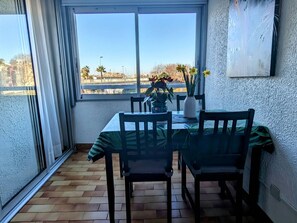 A dining area with a table and chairs, a window with a view of a bridge and trees, and a textured wall. - Studio cabin, parking, cliffs area - 4 guests (Cap d'Agde)