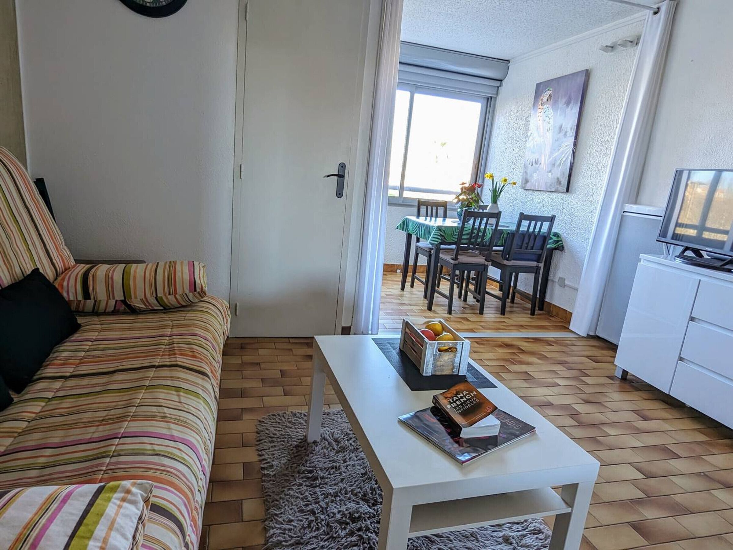 Un salon avec un canapé à rayures, une table basse blanche ornée de livres et de fruits, un coin repas avec une table et des chaises, et un téléviseur posé sur un meuble.
