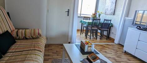 Un salon avec un canapé à rayures, une table basse blanche ornée de livres et de fruits, un coin repas avec une table et des chaises, et un téléviseur posé sur un meuble.