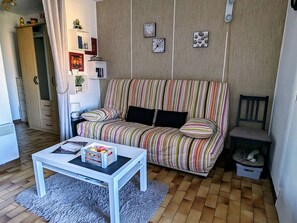A living room with a striped sofa, a white coffee table, a pet bed, and a small bookshelf. - Studio cabin, parking, cliffs area - 4 guests (Cap d'Agde)