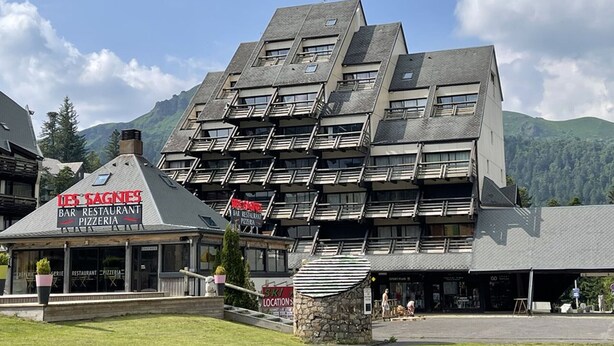 Un edificio de varios pisos con un diseño angular distintivo, que cuenta con balcones y un restaurante llamado "Les Sages" en la planta baja.