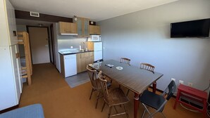 Un espacio habitable compacto con kitchenette, una zona de comedor con mesa y sillas, un televisor montado en la pared y un pequeño taburete rojo.
