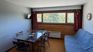 Una habitación con una mesa y sillas de madera, un sofá azul, una ventana con cortinas, un reloj de pared y un radiador.