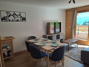A dining area with a table set for four, a television on a wooden cabinet, and a view of the outdoors through a sliding door. - Cozy apartment, mountain view - 6 guests (Valmorel)