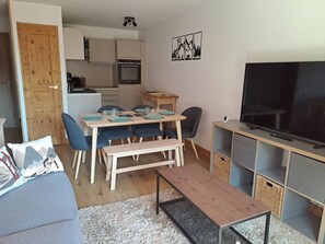 A living room with a sofa, a coffee table, a dining table with chairs, a TV, and a kitchen in the background. - Cozy apartment, mountain view - 6 guests (Valmorel)