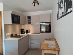 A compact kitchen with a microwave, coffee maker, and oven. There is a small table with a cutting board and a smartphone. - Cozy apartment, mountain view - 6 guests (Valmorel)