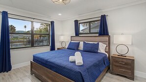 A bedroom with a large bed, two bedside tables, a window with curtains, and a view of the outside. - Canal Front Retreat | Private Pool, Dock, and Game Room (Port Richey)