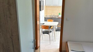 Un pasillo estrecho que da a una cocina con mesa y sillas, una cama con colchón blanco, y una puerta de madera.