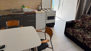 Cocina compacta con estufa, lavamanos y un refrigerador pequeño. Hay una mesa de comedor con sillas, un sofá con un diseño estampado, y vista al exterior a través de una puerta.