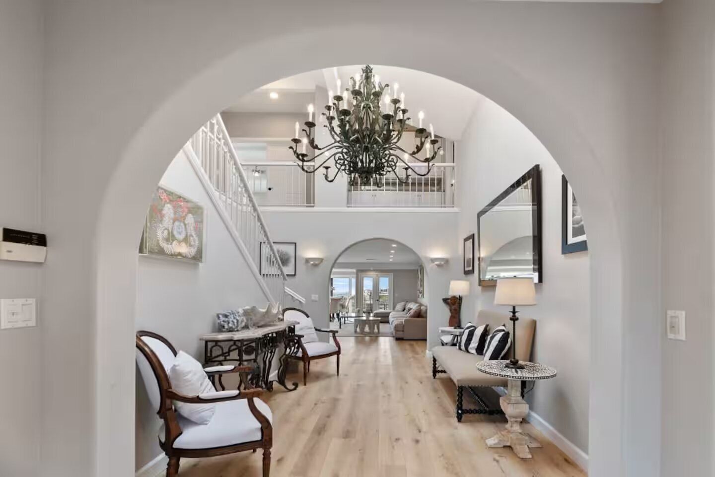A spacious interior with a high ceiling, a grand chandelier, a staircase leading to an upper level, and a dining area with chairs and a table.