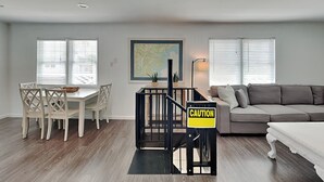A living room with a dining area, a staircase with a caution sign, and a large map on the wall. - 50 Steps to the Beach | Spacious, Gas Grill, Beach Access (Tybee Island)