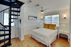 Une chambre avec un lit, un escalier en colimaçon noir, un ventilateur de plafond, une fenêtre avec des stores et un luminaire fixé au mur.
