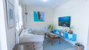 A living room with a sofa, a coffee table, a TV, a fan, and a blue shelving unit with plants. - Charming 2-bedroom Apartment with AC at gorgeous Crane Beach (Bridgetown)
