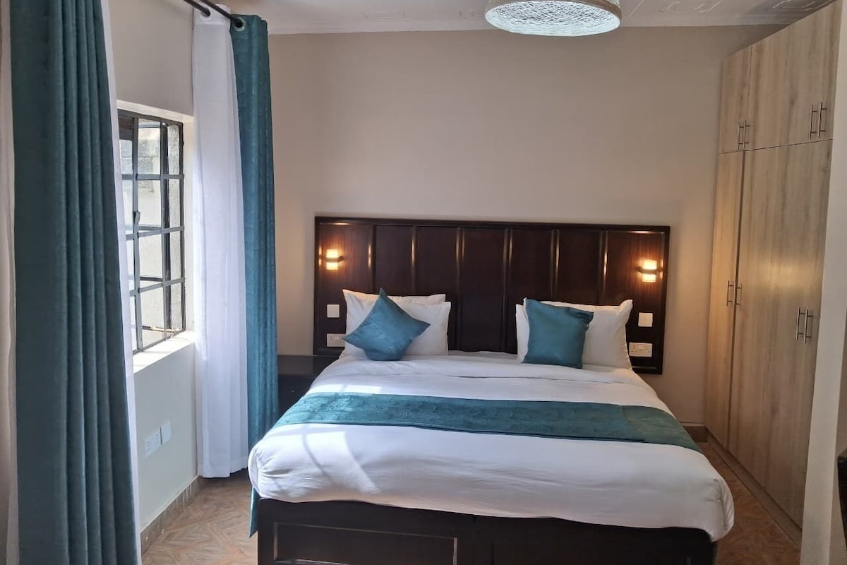 A hotel room with a bed, a window with curtains, a headboard with wall-mounted lights, and a wardrobe.