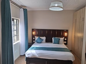A hotel room with a bed, a window with curtains, a headboard with wall-mounted lights, and a wardrobe. - Riata Loltome (Nyahururu)