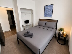 A bedroom with a bed, a bedside table with a lamp, a TV on a stand, and a door leading to another room.