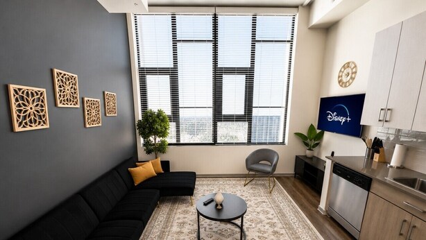 Een moderne woonkamer met een zwarte modulaire bank, een ronde salontafel, een televisie en een keuken met een vaatwasser.