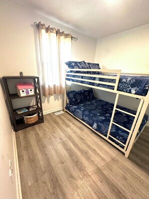 A bunk bed with a patterned bedspread, a small shelf unit with a house-shaped object, and a window with curtains. - House in Panama City Beach ~ Mermaid Oasis (Panama City)