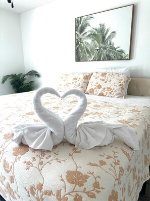 Ein Hotelzimmer mit einem Bett, das eine blumige Tagesdecke, eine herzförmige Handtuchgestaltung, ein Palmen-Gemälde an der Wand und eine Topfpflanze aufweist.