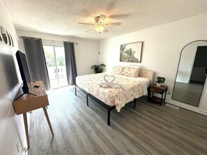 Ein Schlafzimmer mit einem Bett, einem Deckenventilator, einem Fenster mit Vorhängen, einem Nachttisch mit Pflanze und einem Spiegel.