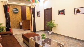 A hotel lobby with a reception desk, a sofa, a glass-top table with a plant, and framed pictures on the walls.