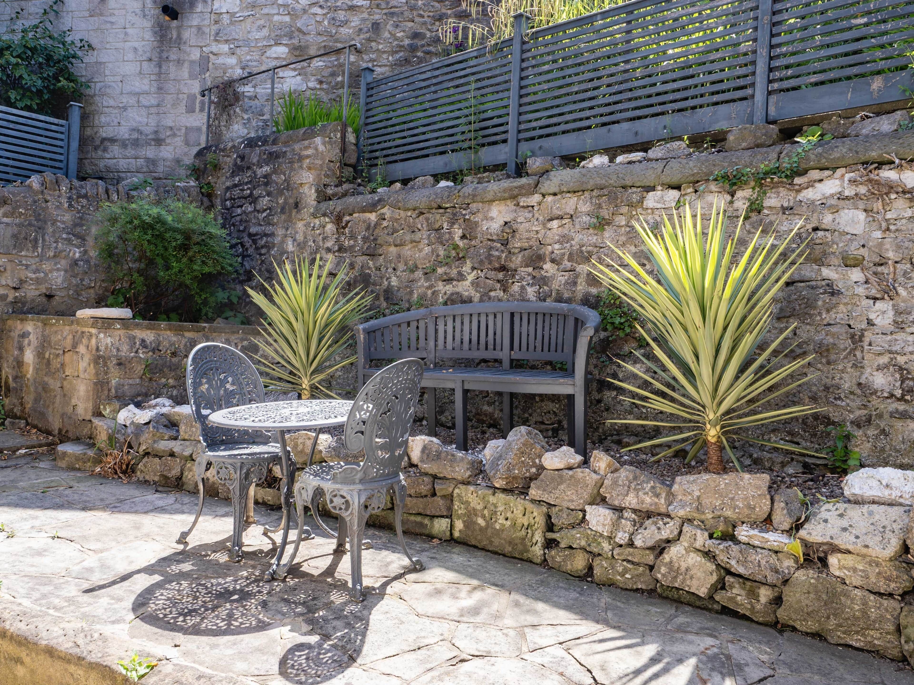 A stone wall with a bench and two metal chairs, surrounded by plants.