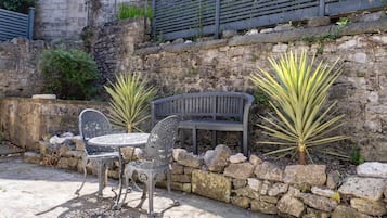 A stone wall with a bench and two metal chairs, surrounded by plants.