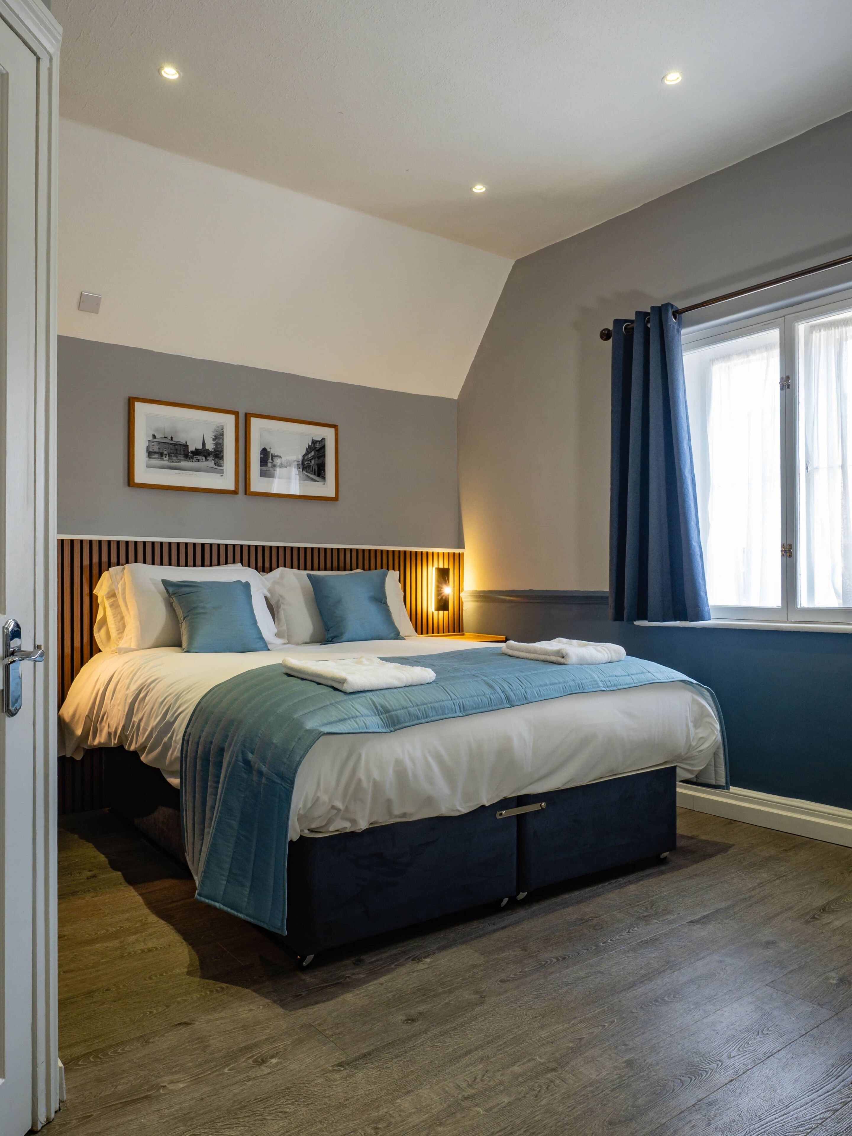 A bedroom with a bed, a window with curtains, a wall-mounted light, and two framed pictures.