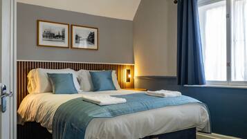 A bedroom with a bed, a window with curtains, a wall-mounted light, and two framed pictures.