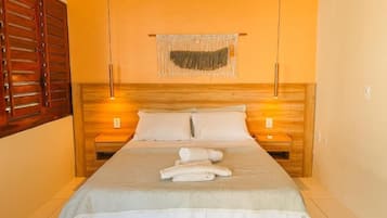 Habitación de hotel con cama, cabecero de madera, dos mesitas de noche, un macramé en la pared y una puerta de madera.
